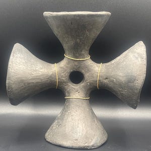 May include: A dark gray, cross-shaped sculpture with a central circular opening. The sculpture has four flared arms and is adorned with thin, gold-colored wire accents. The piece is likely made of ceramic or a similar material.