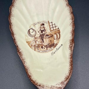 May include: Cream-colored, shell-shaped ceramic dish with a brown scalloped edge. The center features a sepia-toned illustration of a cooper working on a barrel, with the words "Le Tonnellier" and "Bordeaux".