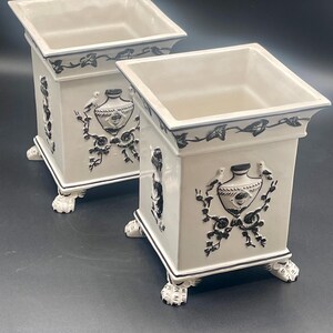 May include: Two square off-white ceramic planters featuring black relief designs of urns, birds, and vines. Each planter has a flared rim, a smooth interior, and four decorative feet shaped like animal paws with detailed claw markings.