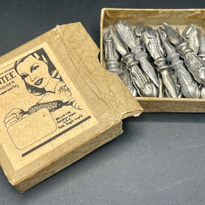 Könnte beinhalten: Ein Set aus silberfarbenen Maiskolbenhaltern in einer beigen Pappschachtel. Der Deckel zeigt eine schwarz-weiße Illustration einer Person, die einen Maiskolben hält, mit dem Text "GENUINE SILVA KROME DAINTEE CORN HOLDER Convenient and Tidy".