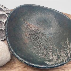 May include: A handmade, oval ceramic bowl in teal and blue hues. The bowl has an embossed botanical design in a lighter colour. The bowl is approximately 25 cm long and 20 cm wide, suitable for serving or display.