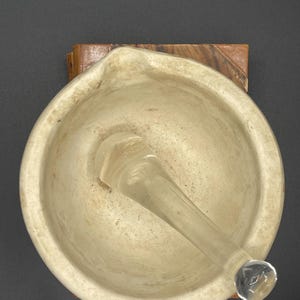 May include: A mortar and pestle set. The mortar is a light beige color with a rounded shape and a pouring spout. The pestle is clear glass. The set is on a wooden base, against a dark background.