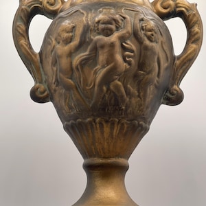 May include: A bronze-colored decorative urn with two ornate handles. The urn features a relief of dancing figures around the body. The base and handles have detailed, textured designs. The overall style is classical.