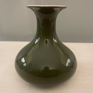 Może przedstawiać: Ciemnozielona ceramiczna waza o pękatym korpusie i smukłej, rozszerzającej się szyi. Waza ma błyszczące wykończenie i kremowy brzeg. Waza stoi na jasnej powierzchni.