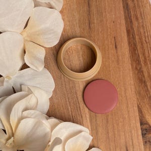 Può includere: Un tagliapasta circolare color beige con un pezzo rotondo e pieno abbinato. Gli oggetti sono su una superficie di legno, con petali di fiori bianchi sullo sfondo. Il tagliapasta ha la forma di un anello.