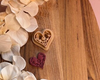 May include: A wooden cutting board with a heart-shaped cookie cutter and a heart-shaped cookie. The cookie cutter is beige and the cookie is dark red. White and cream-colored flowers are in the background.
