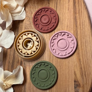 May include: Four circular cookie cutters in shades of brown, pink, and green, with a floral design. One is a tan-colored cutter, and the others are solid-colored impressions. The cutters are arranged on a wooden cutting board.