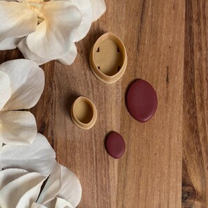 May include: A collection of beige and burgundy polymer clay cutters and shapes on a wooden surface. The cutters are in oval and teardrop shapes, with a larger oval shape and two smaller oval shapes. White flower petals are in the background.