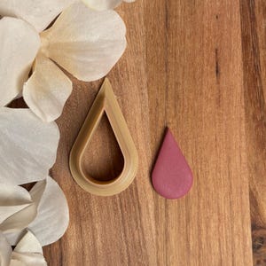 May include: A teardrop-shaped clay cutter and a small, pink teardrop-shaped clay piece sit on a wooden surface. The cutter is beige, and the clay piece is a solid, muted pink. White flower petals are in the background.