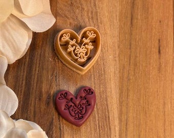 May include: Two heart-shaped cookie cutters. One is tan and the other is burgundy. Both have a floral design in the center. The cutters are on a wooden surface with white flower petals in the background.