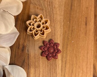 May include: Two flower-shaped cookie cutters. One is a light tan color, and the other is a deep red. The cutters are on a wooden surface, with white and cream-colored flower petals in the background. The cutters have detailed petal designs.