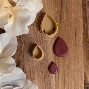May include: Three teardrop-shaped items on a wooden surface. Two are tan-colored cookie cutters, and the other two are solid maroon teardrops. The teardrops vary in size. White flower petals are in the upper left corner.