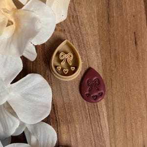 May include: A beige teardrop-shaped cookie cutter with a bow and three hearts, next to a maroon teardrop-shaped cookie with the same design. The items are on a wooden surface, with white flowers in the background.