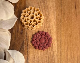 May include: Two flower-shaped objects on a wooden surface. One is a light tan color with a detailed, open design. The other is a solid, maroon-colored flower. White flower petals are in the background.