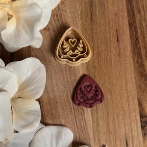 May include: Two heart-shaped cookie cutters with a heart and leaf design. One is beige, the other is maroon. The cutters are on a wooden surface, with white flower petals in the background.