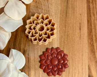 May include: Two flower-shaped cookie cutters. One is a light tan color, and the other is a deep reddish-brown. The cutters are made of plastic and have detailed petal designs. The background is a wooden surface.