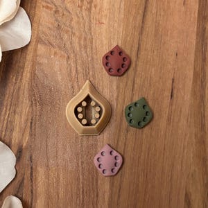 May include: Four teardrop-shaped cookie cutters in shades of brown, red, green, and pink. Each cutter has a series of small, circular cutouts. The largest cutter has a teardrop shape with a hollow center and circular cutouts.