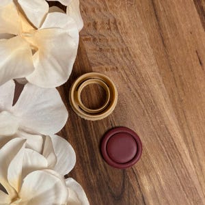 Può includere: Un sigillo di cera bordeaux e due formine per biscotti beige sono su una superficie di legno, con petali di fiori bianchi sullo sfondo. Il sigillo è ovale e le formine sono circolari. La superficie di legno ha venature naturali.