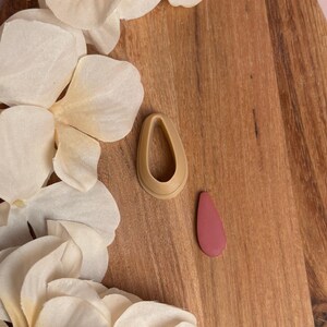 May include: A teardrop-shaped earring and a matching earring cutter in a light brown color, next to a solid pink earring. The earrings are on a wooden surface, with white flower petals in the background.