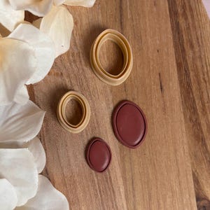 May include: An overhead shot displays four oval-shaped polymer clay cutters in varying sizes. Two are tan, and two are a deep red-brown. The cutters are arranged on a wooden surface, with white flower petals in the background.