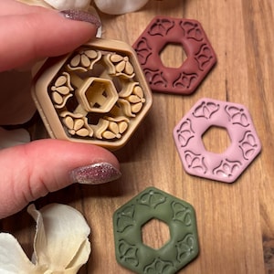 Può includere: Un tagliapasta esagonale beige in argilla polimerica con un motivo floreale è tenuto vicino a tre pezzi di argilla finiti in rosso, rosa e verde. I pezzi di argilla hanno un motivo floreale simile e un foro esagonale al centro.