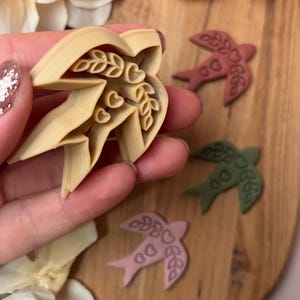 May include: A beige cookie cutter in the shape of a flower with leaf and heart details is held in a hand. Three bird-shaped cookies in shades of pink, green, and red are on a wooden surface. The cookies have leaf and heart details.