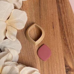 May include: A tan-colored teardrop-shaped cookie cutter and a matching mauve-colored clay earring component rest on a wooden surface. White flower petals are scattered around the objects.
