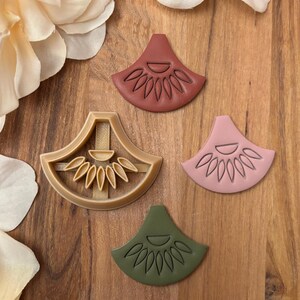 Könnte beinhalten: Eine Auswahl an fächerförmigen Tonstücken und ein passender Ausstecher. Die Tonstücke sind in den Farbtönen Rosa, Grün und Braun gehalten, jedes mit einem detaillierten floralen Design. Der Ausstecher ist beige.