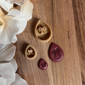 May include: A collection of teardrop-shaped cookie cutters and embossed cookies. The cutters are beige, and the cookies are a deep burgundy color. Each piece features a bow design. The items are arranged on a wooden surface.