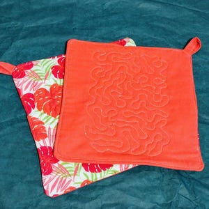 May include: Two square pot holders. One is a vibrant coral color with a quilted design. The other has a white background with a tropical floral pattern of red flowers and green leaves. Both have a small loop for hanging.