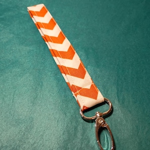 May include: A fabric wrist strap with a white and orange chevron pattern. The strap is attached to a silver-colored metal clasp. The strap is approximately 8 inches long.