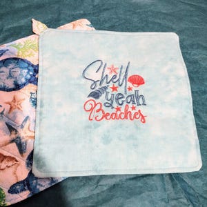May include: Two square pot holders. One has a blue sea turtle and starfish pattern. The other is light blue with the embroidered phrase "Shell Yeah Beaches" in blue and red, with seashell and starfish accents.