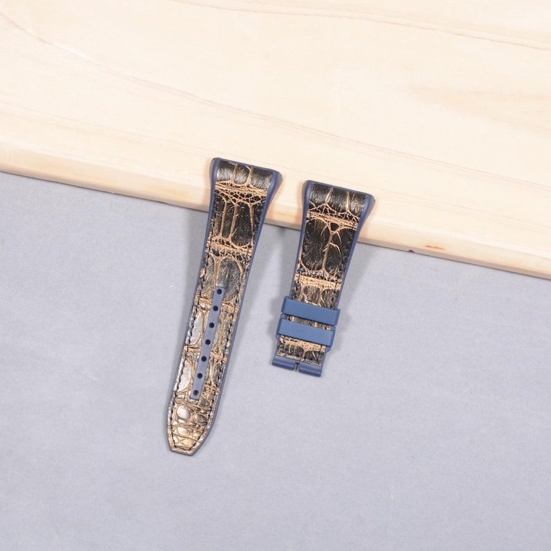 K&ouml;nnte beinhalten: Zweiteiliges Uhrenarmband in Blau und Gold. Das Armband hat ein strukturiertes, krokodilartiges Muster in Gold- und Braunt&ouml;nen. Das Armband hat mehrere L&ouml;cher zur Anpassung und eine blaue Schnalle.