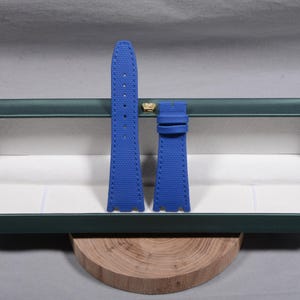 Puede incluir: Un par de correas de reloj azul brillante con una superficie texturizada y costuras blancas. Las correas se muestran dentro de una caja verde con un emblema dorado.