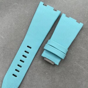 May include: A light blue watch strap with a textured surface. The strap has a series of evenly spaced holes for adjustment and a metal clasp. The strap is designed to attach to a watch face.
