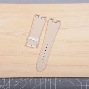 May include: Two beige leather watch straps with silver-tone metal clasps. One strap has a white leather buckle. The straps are on a light wood surface.