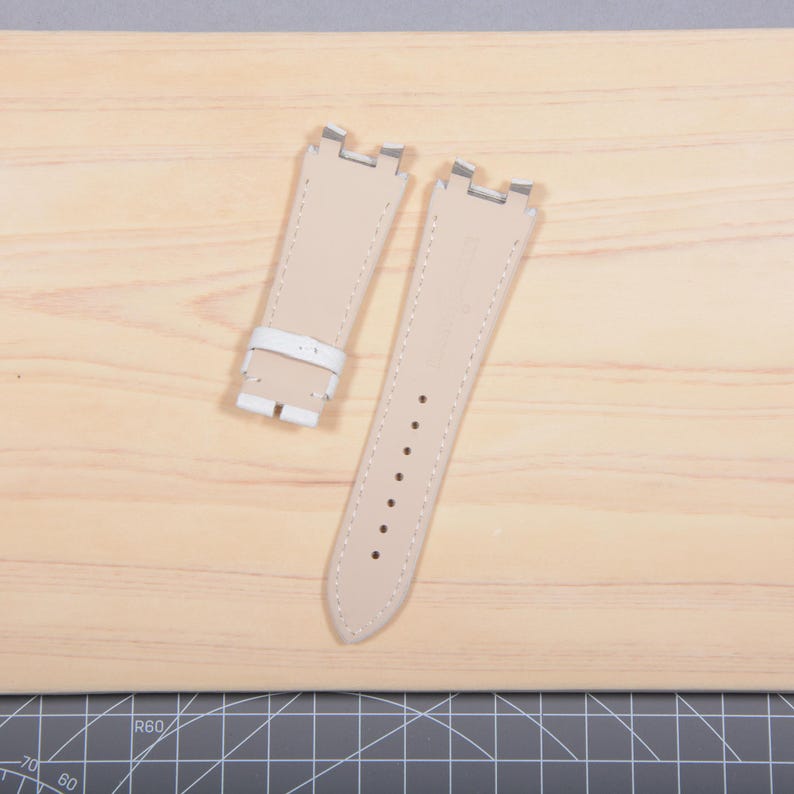 May include: Two beige leather watch straps with silver-tone metal clasps. One strap has a white leather buckle. The straps are on a light wood surface.
