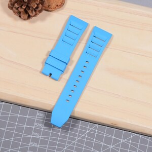 Op de afbeelding: Tweedelig lichtblauwe horlogeband. De band heeft rechthoekige uitsparingen aan &eacute;&eacute;n uiteinde en een reeks kleine gaatjes aan het andere. De band is gemaakt van flexibel materiaal en is ontworpen om aan een horlogekast te bevestigen.