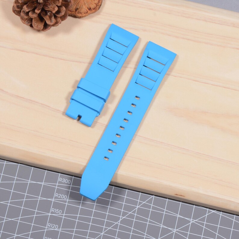 Op de afbeelding: Tweedelig lichtblauwe horlogeband. De band heeft rechthoekige uitsparingen aan &eacute;&eacute;n uiteinde en een reeks kleine gaatjes aan het andere. De band is gemaakt van flexibel materiaal en is ontworpen om aan een horlogekast te bevestigen.
