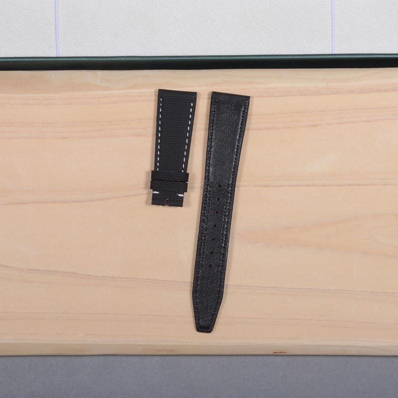 May include: Two-piece black watch strap with white stitching. The shorter piece has a rectangular shape with a buckle, while the longer piece is tapered with multiple holes for adjustment. The strap is made of a textured material.