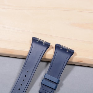 K&ouml;nnte beinhalten: Zwei marineblaue Uhrenarmb&auml;nder mit einem gen&auml;hten Design und einer Schnalle. Die Armb&auml;nder bestehen aus einem glatten Material und sind zur Befestigung an einem Zifferblatt konzipiert. Die Armb&auml;nder werden auf einer hellbraunen Holzoberfl&auml;che pr&auml;sentiert.