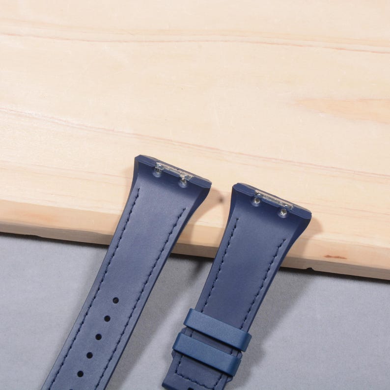 K&ouml;nnte beinhalten: Zwei marineblaue Uhrenarmb&auml;nder mit einem gen&auml;hten Design und einer Schnalle. Die Armb&auml;nder bestehen aus einem glatten Material und sind zur Befestigung an einem Zifferblatt konzipiert. Die Armb&auml;nder werden auf einer hellbraunen Holzoberfl&auml;che pr&auml;sentiert.