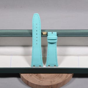 Puede incluir: Correa de reloj de dos piezas en color turquesa claro. Las correas est&aacute;n hechas de un material texturizado y presentan costuras a lo largo de los bordes. Se exhiben en una caja verde con un emblema dorado.