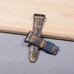 K&ouml;nnte beinhalten: Zwei Uhrenarmb&auml;nder mit Krokodilhautmuster in Gold und Dunkelblau. Die Armb&auml;nder haben eine dunkelblaue Schnalle und liegen auf einer hellen Holzoberfl&auml;che.