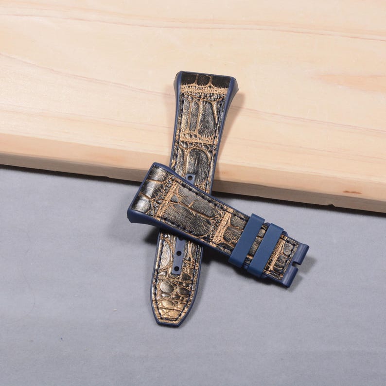 K&ouml;nnte beinhalten: Zwei Uhrenarmb&auml;nder mit Krokodilhautmuster in Gold und Dunkelblau. Die Armb&auml;nder haben eine dunkelblaue Schnalle und liegen auf einer hellen Holzoberfl&auml;che.