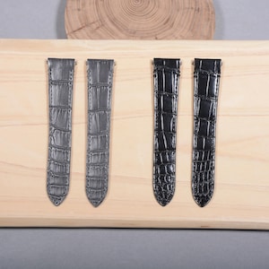 Puede incluir: Cuatro correas de reloj, dos grises y dos negras, se muestran sobre una superficie de madera clara. Las correas tienen un patrón texturizado similar al de un cocodrilo y están diseñadas para sujetarse a la esfera del reloj.