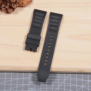 Op de afbeelding: Tweedelig zwart horlogebandje. De band heeft een gestructureerd ontwerp met horizontale ribbels en een reeks gaten voor aanpassing. De horlogeband is gemaakt van een flexibel materiaal en is ontworpen om aan een horlogekast te worden bevestigd.