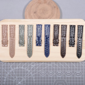 Pode incluir: Uma coleção de pulseiras de relógio em várias cores, incluindo bege, cinza, azul, marrom e preto. As pulseiras estão dispostas em uma tábua de madeira e apresentam um design texturizado. O fundo inclui uma fatia de madeira e uma esteira de corte cinza.