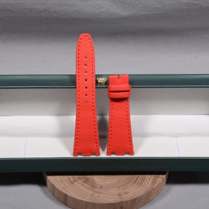 Puede incluir: Juego de correas de reloj de color rojo brillante. Las correas est&aacute;n hechas de un material texturizado y presentan costuras a lo largo de los bordes. La correa tiene varios agujeros para ajustar y una hebilla. El juego se muestra en una caja verde y blanca.