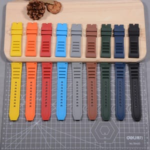 Op de afbeelding: Een verzameling horlogebandjes in diverse kleuren, waaronder geel, oranje, rood, blauw, grijs, bruin, groen, marineblauw en zwart. De bandjes zijn gemaakt van flexibel materiaal en hebben een gestructureerd ontwerp met meerdere gaten voor aanpassing.